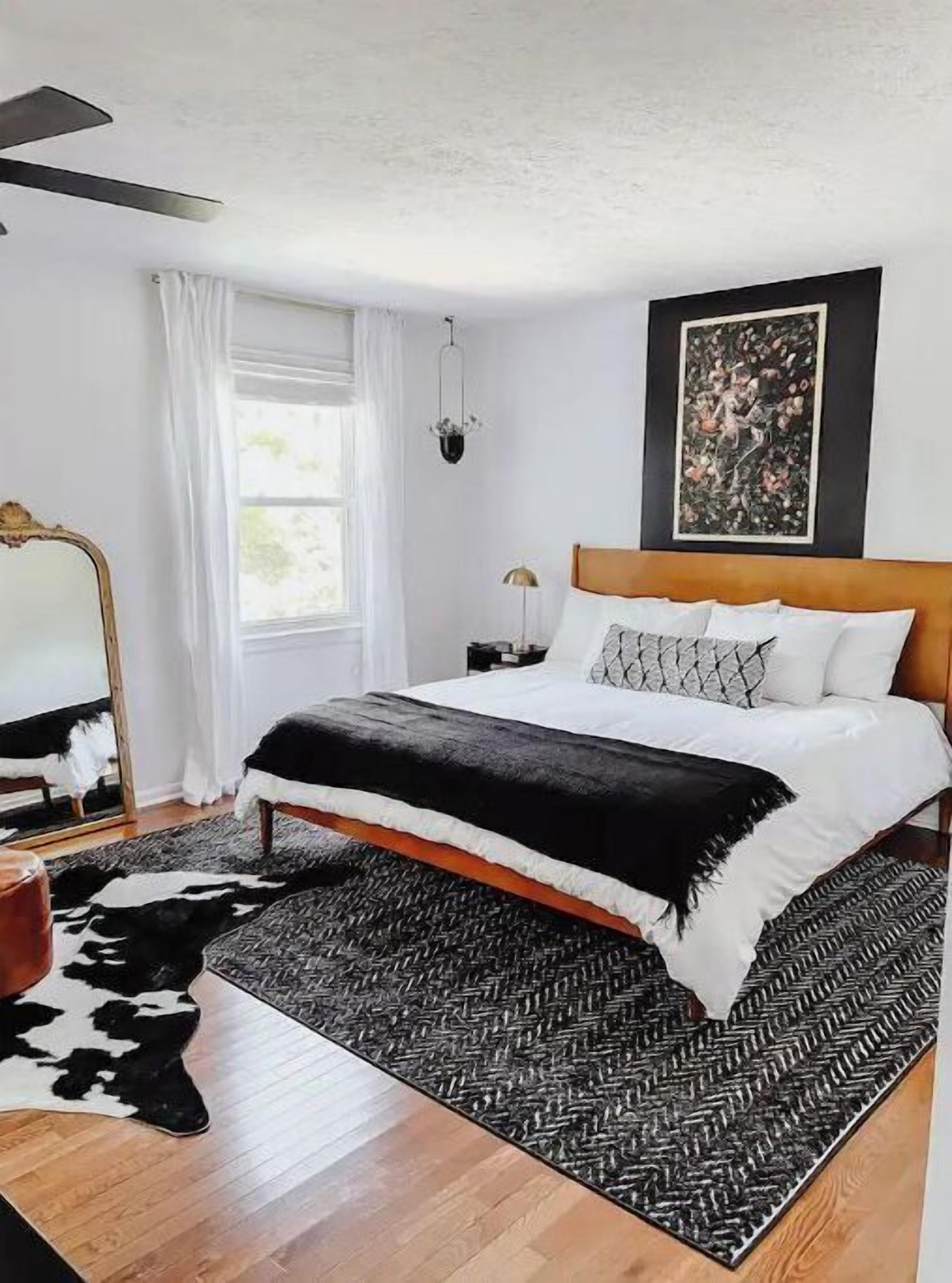 A bedroom with a bed, a mirror, a window with curtains, a hanging light, and a wall-mounted painting.