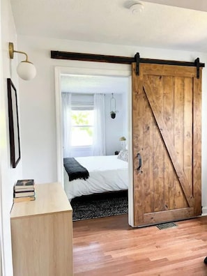 Un dormitorio con puerta corredera de madera, una cama con ropa de cama blanca, una mesita de noche con libros y una lámpara colgante.