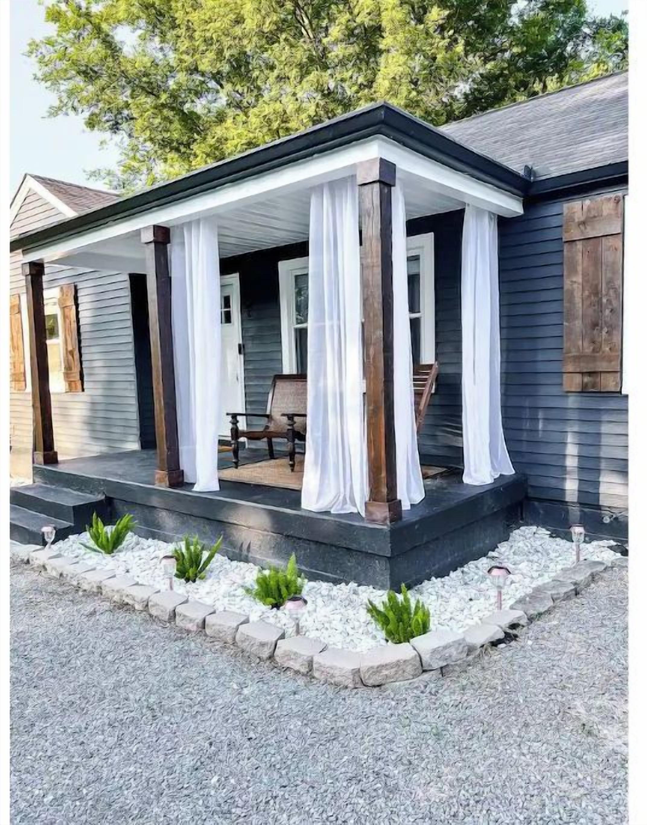 Une maison avec un porche couvert, des rideaux blancs et une allée en gravier.