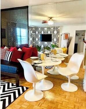 A modern living room with a dining area, featuring a sofa with red cushions, a patterned wall, and a ceiling fan.