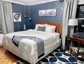 A neatly made bed with a white and gray blanket, a wooden headboard, and a cityscape painting on the wall.