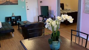 A dining area with a wooden table, chairs, and a vase of white flowers. There is a television on a stand and a door leading to another room.