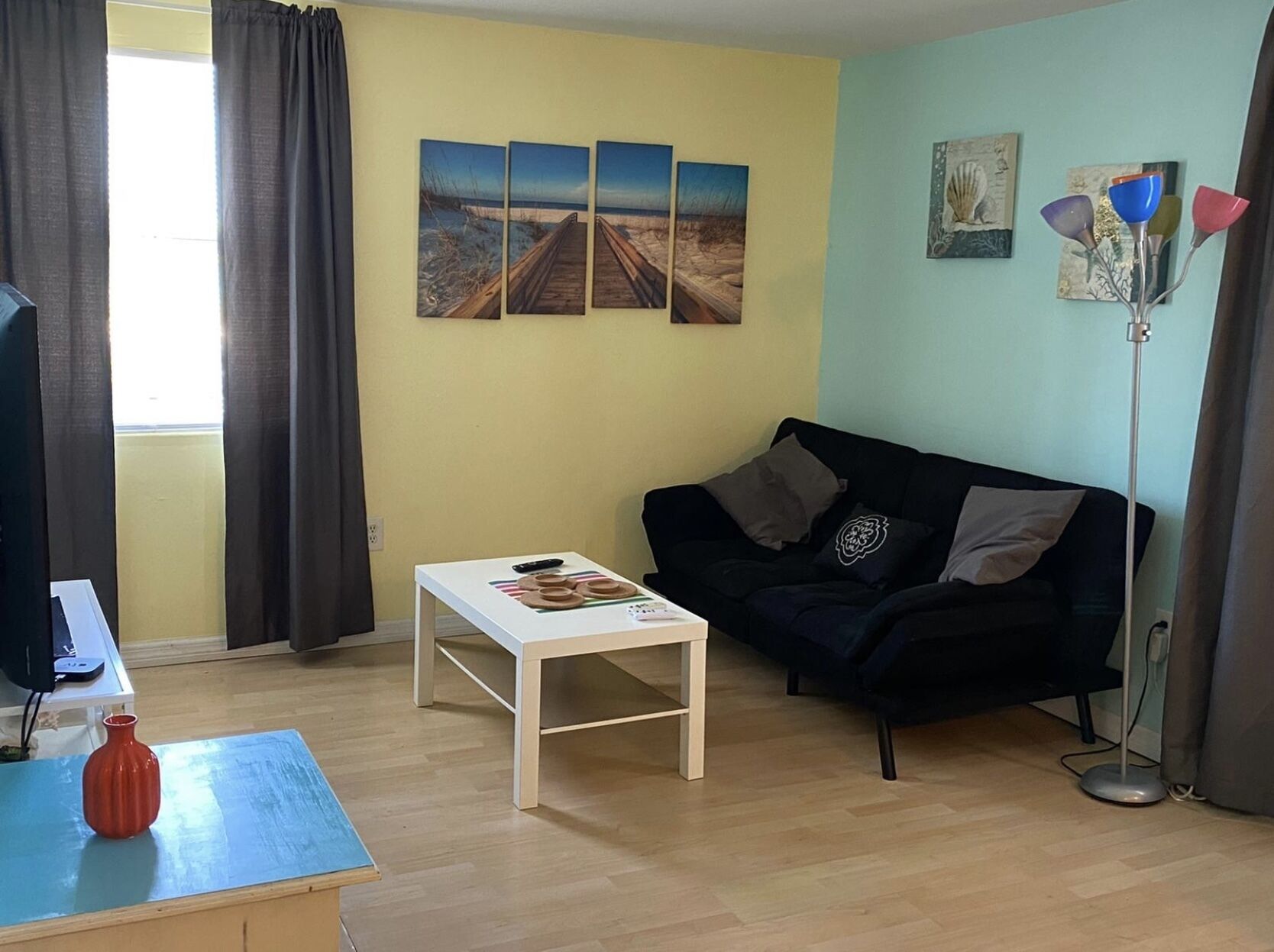 A living room with a wooden floor, a black sofa, a white coffee table, and a TV on a stand. There are three paintings on the wall and a floor lamp.