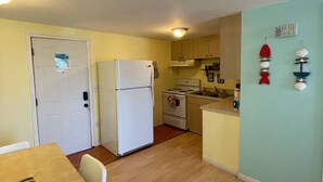 A compact kitchen with a refrigerator, stove, and sink. There is a dining table with chairs and a small island. The walls are painted in light colors, and there is a decorative sign on the wall. - 1 BR - ocean view - unit 2 (Fort Myers Beach)
