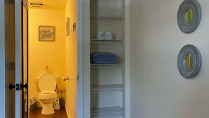 A hallway leading to a bathroom with a toilet, a shelf with toiletries, and a door with a doormat. - 1 BR - ocean view - unit 2 (Fort Myers Beach)