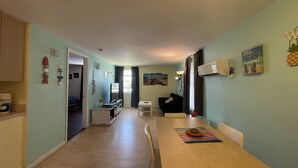 A spacious living area with a long wooden table, a sofa, and a TV. There are decorative items on the walls and a window with curtains. - 1 BR - ocean view - unit 2 (Fort Myers Beach)