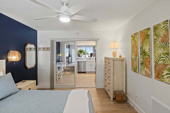 Quarto com cama, mesa de cabeceira, abajur, ventilador de teto e vista para a cozinha.