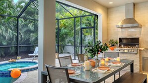 An outdoor dining area with a pool, a grill, and a view of lush greenery.