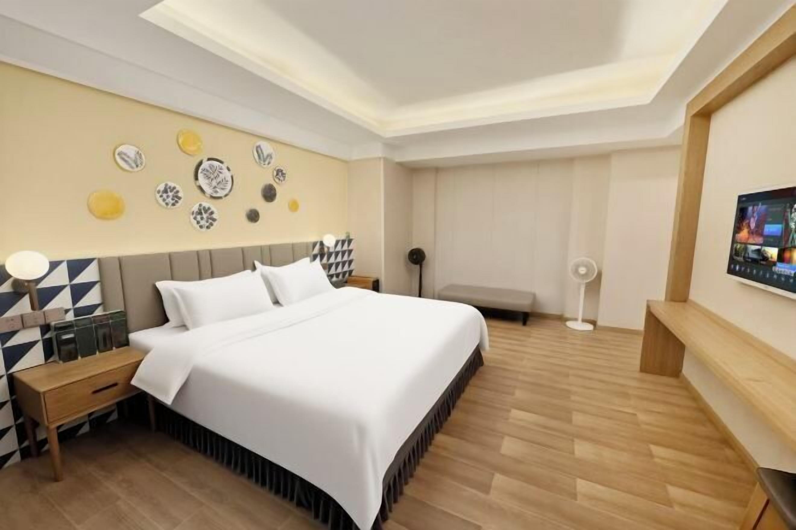 A modern hotel room with a large bed, a bedside table, a wall-mounted TV, and a ceiling light fixture.