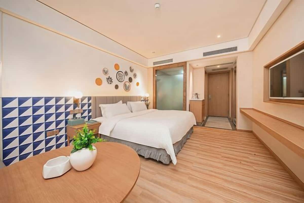 A modern hotel room with a bed, bedside tables, a round table with a plant, a wall with geometric patterns, and a large window.