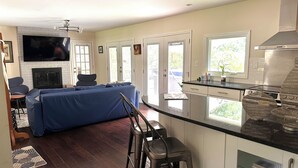 A modern kitchen with white cabinets, a black countertop, and a stainless steel range hood. There is a blue sofa and a dining area with a table and chairs. - Beautiful Island Home (Savannah)