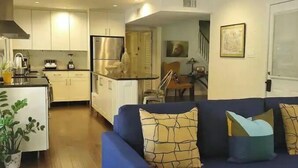 A modern living room with a blue sofa, a kitchen in the background, and a staircase leading to another level. - Beautiful Island Home (Savannah)