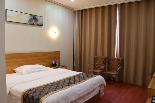 A hotel room with a wooden headboard, a bed with white linens, a patterned throw blanket, a nightstand with a phone, two wicker chairs, a desk, and a large window with curtains. - Jinjiang Zhixing Hotel (Fengshan)