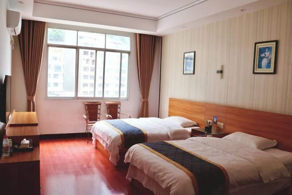 A hotel room with two beds, a wooden headboard, a window with curtains, a desk with a lamp, and a chair.