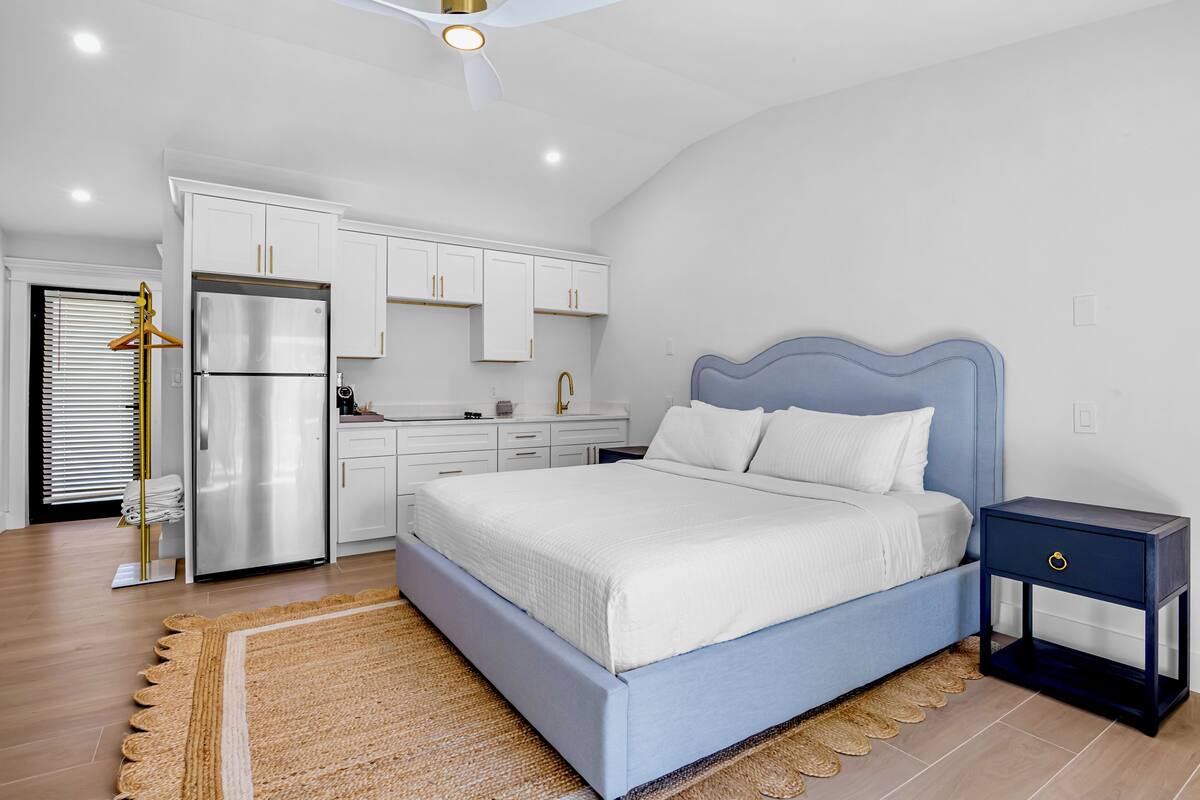 Un dormitorio con cama, mesita de noche y una zona de cocina con armarios y un refrigerador.