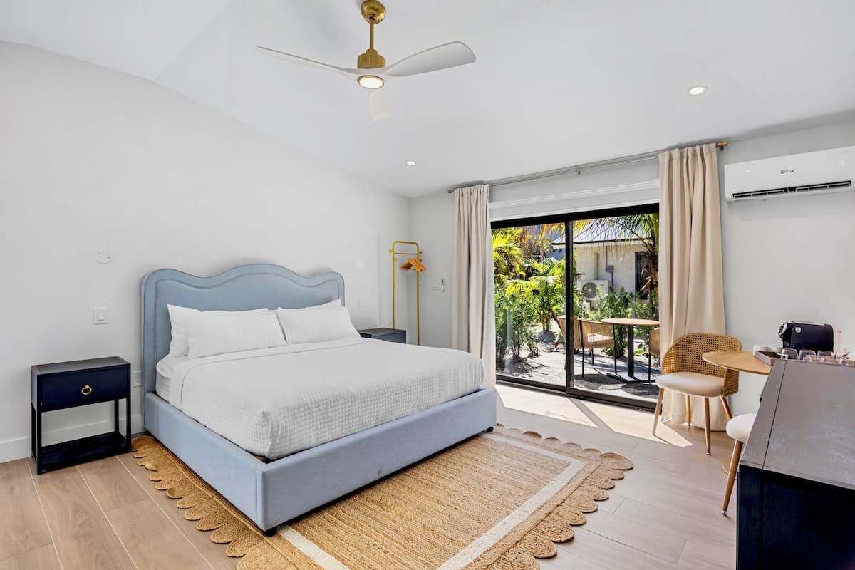 Un dormitorio con cama, mesitas de noche, ventilador de techo, ventana grande con cortinas y vistas al exterior.