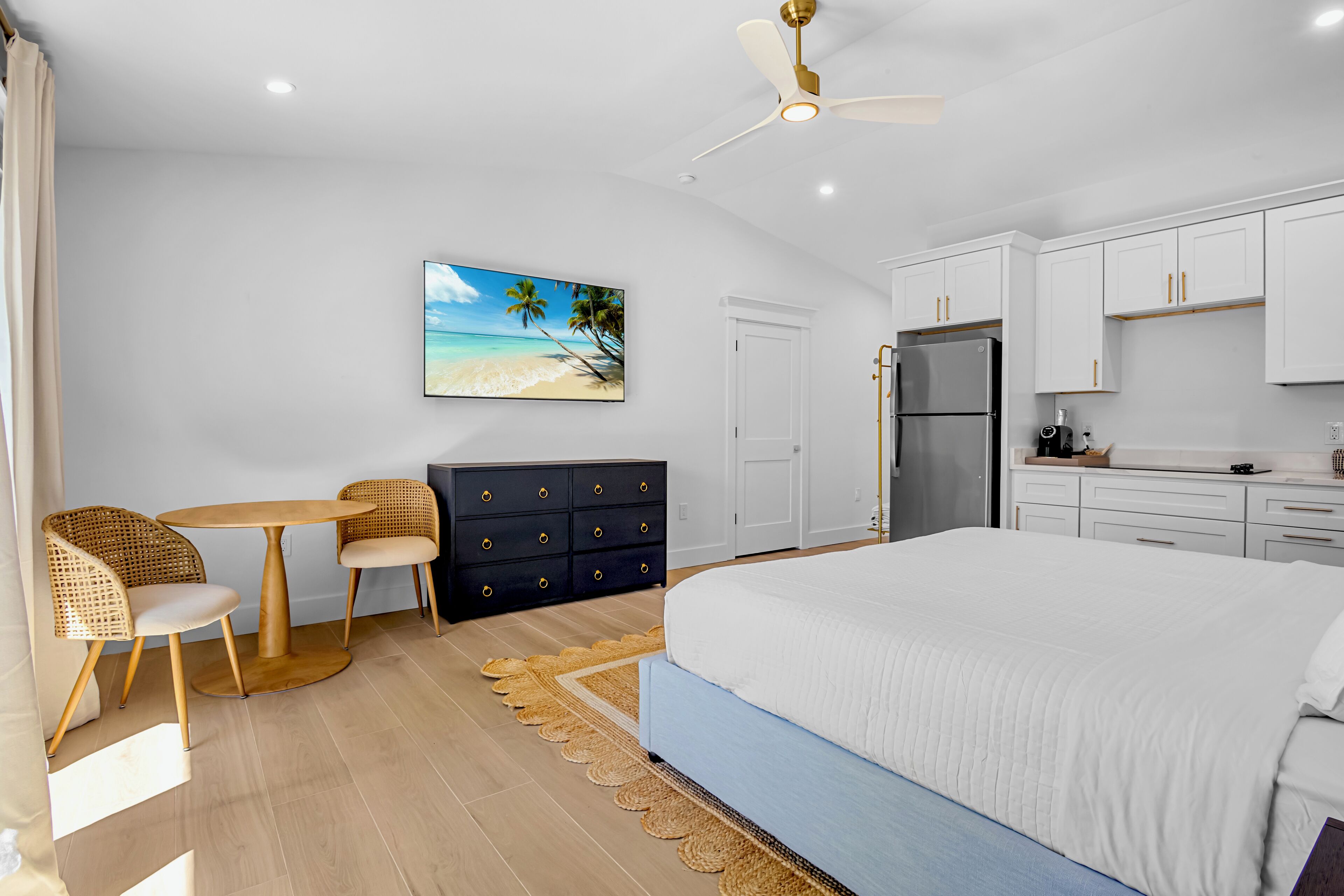 A bedroom with a bed, a desk, chairs, a dresser, a refrigerator, and a ceiling fan.