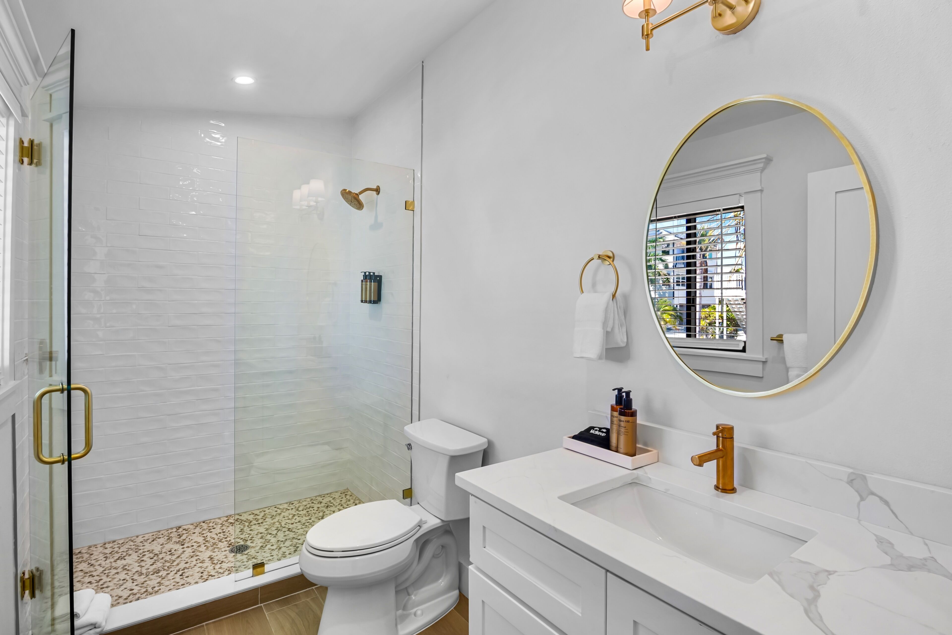 Un baño moderno con ducha de vidrio, inodoro blanco, lavamanos de mármol y un espejo grande con marco dorado.