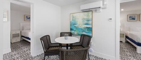 A hotel room with a white bed, a small table with chairs, a large abstract painting, and an air conditioning unit on the wall.