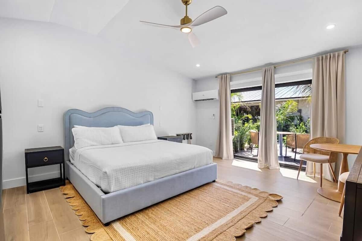 Un dormitorio con cama, mesita de noche, ventilador de techo, ventana con cortinas y balcón con sillas.