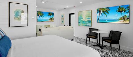 A hotel room with a large bed, a desk, two chairs, a small table, and artwork on the walls.