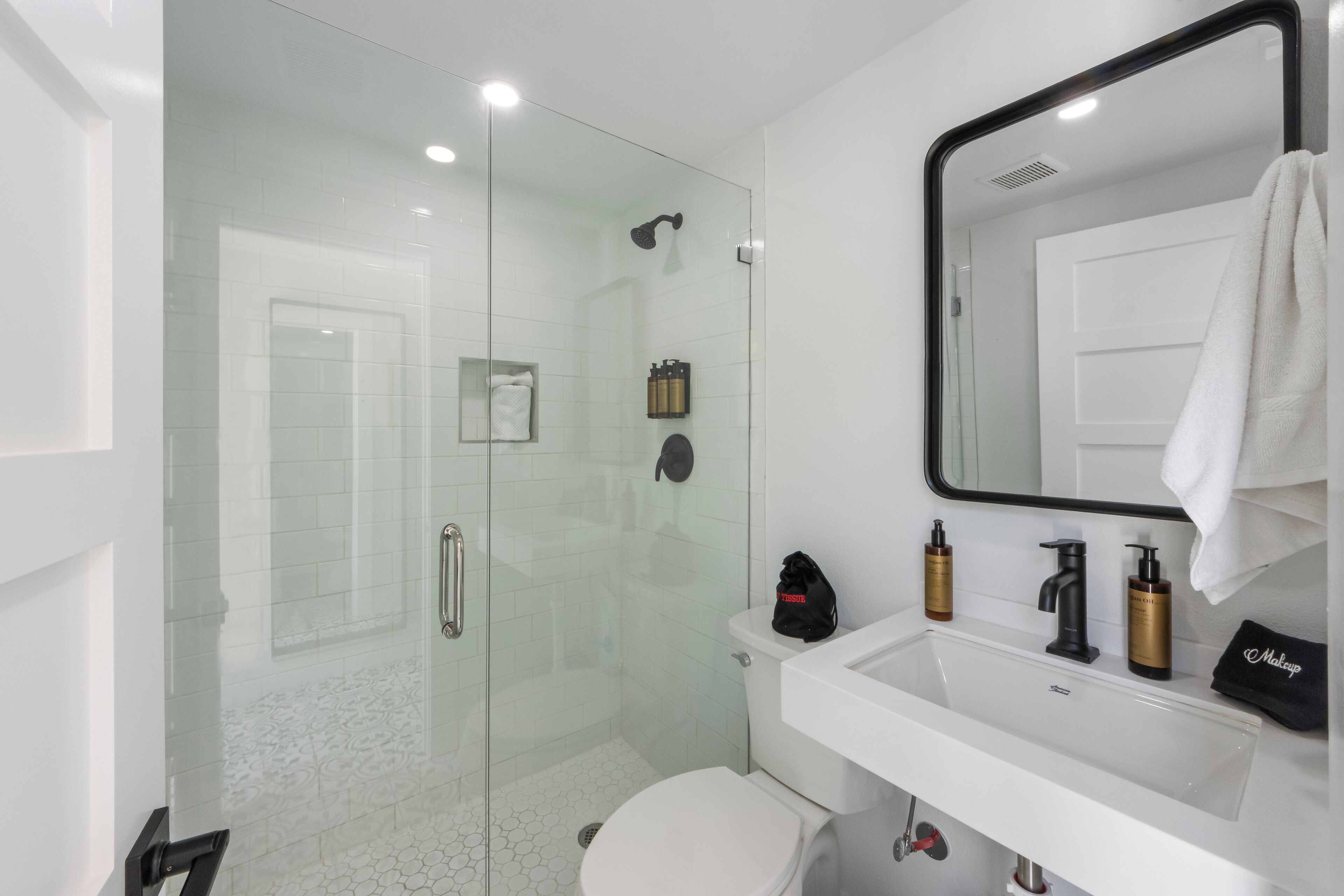 A modern bathroom with a glass-enclosed shower, a white sink with toiletries, and a large mirror.