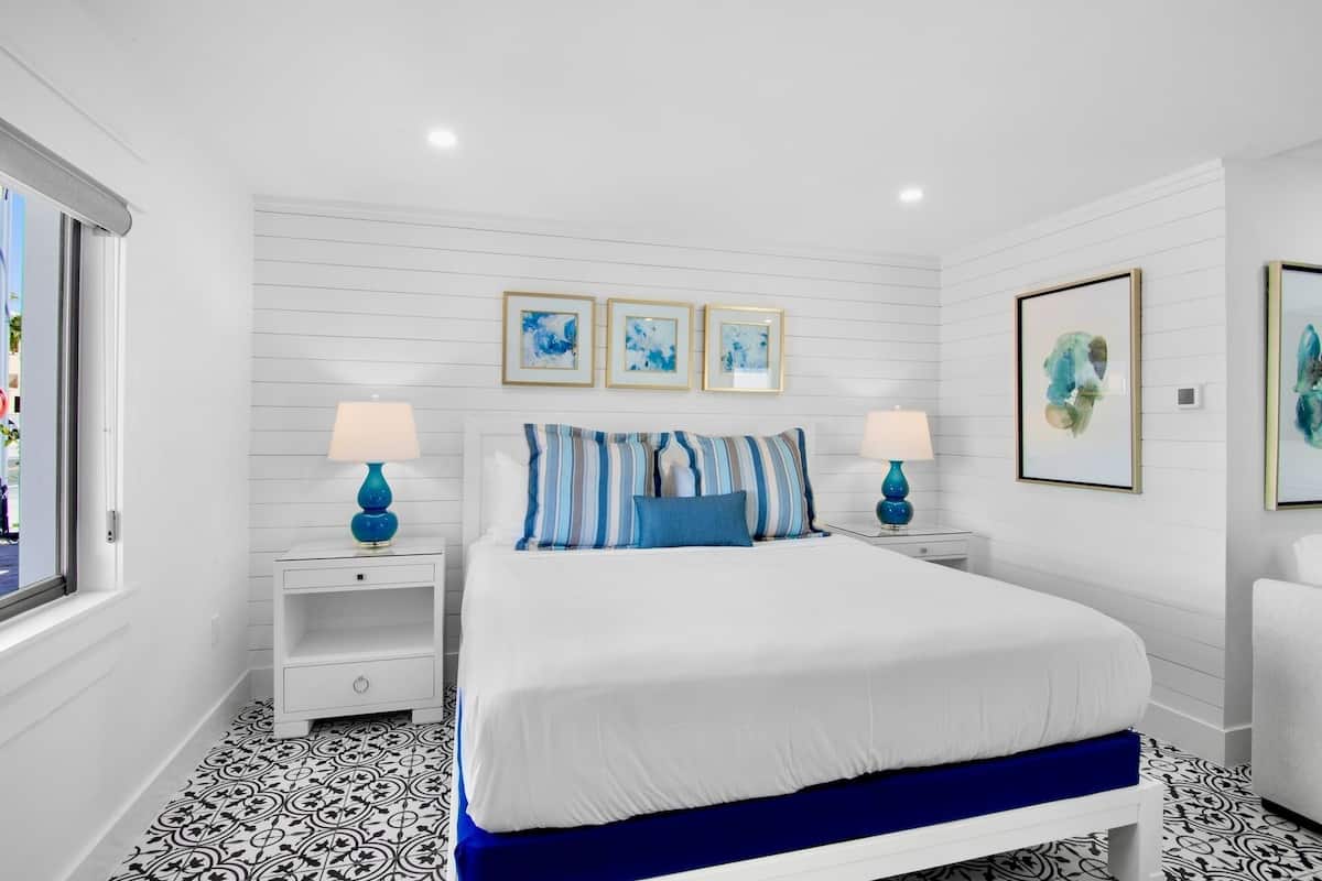 Un dormitorio con una cama blanca, almohadas a rayas azules y blancas, dos mesitas de noche con lámparas y tres cuadros enmarcados en la pared.