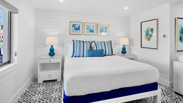 A bedroom with a white bed, blue and white striped pillows, two bedside tables with lamps, and three framed artworks on the wall.