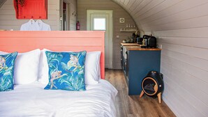 Une chambre confortable avec un lit, une étagère rouge contenant des livres et une plante, un banc en bois et un petit coin cuisine équipé d’une machine à café.