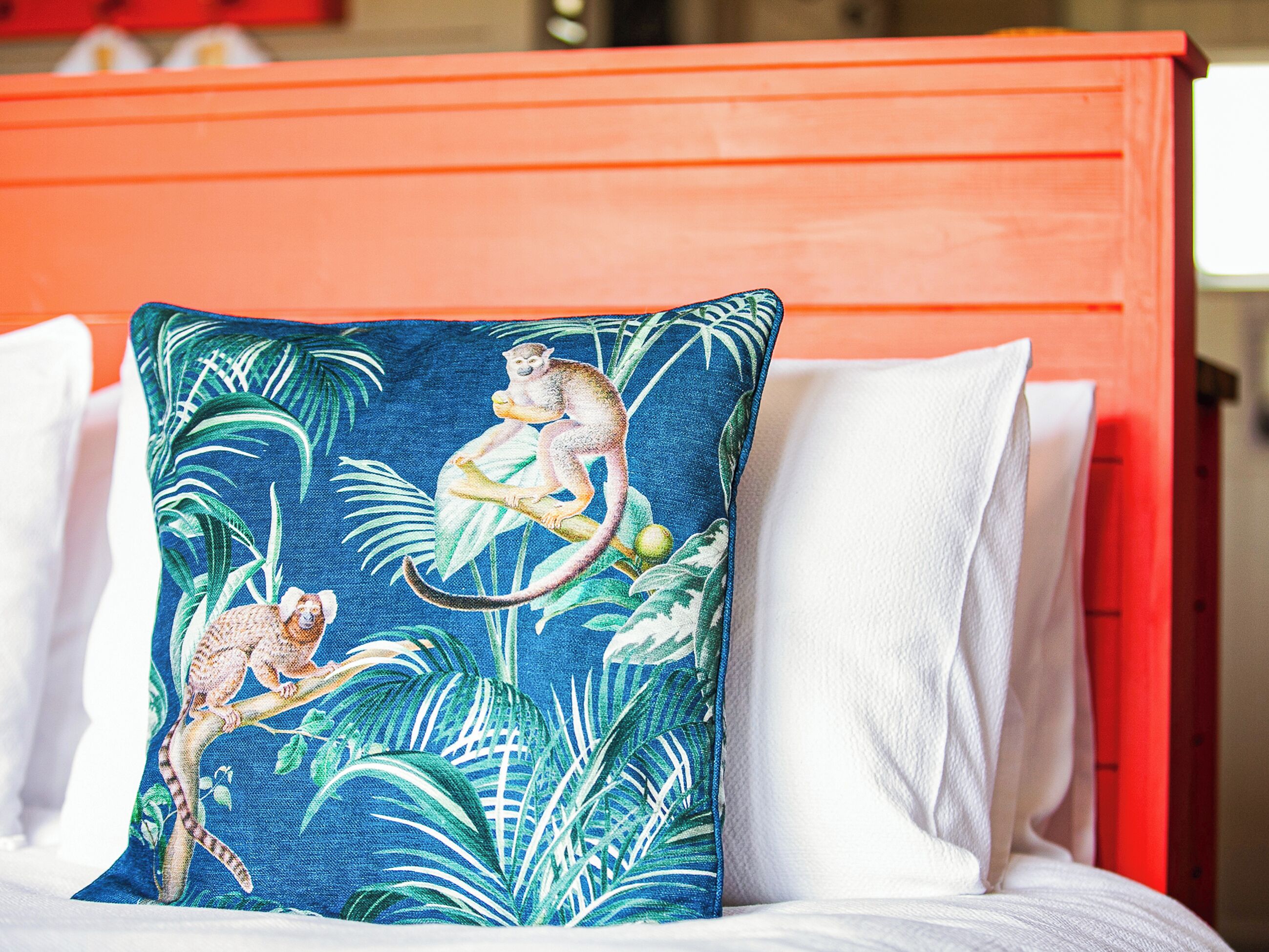 Una cama con almohadas blancas y una sola almohada decorativa con un mono y hojas tropicales.