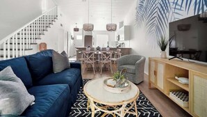 A modern living room with a blue sofa, a round coffee table, a grey armchair, a wooden TV stand, and a dining area in the background. - Beach Getaway - Team Joseph Ellen (Atlantic Beach)