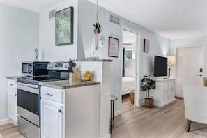 A modern kitchen with white cabinetry, a built-in oven, and a microwave. There is a small island with a sink and a bowl of lemons. The room has light wooden flooring and white walls.