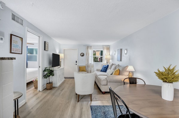 A modern living room with a sofa, coffee table, and a TV. There is a dining table with chairs and a potted plant.