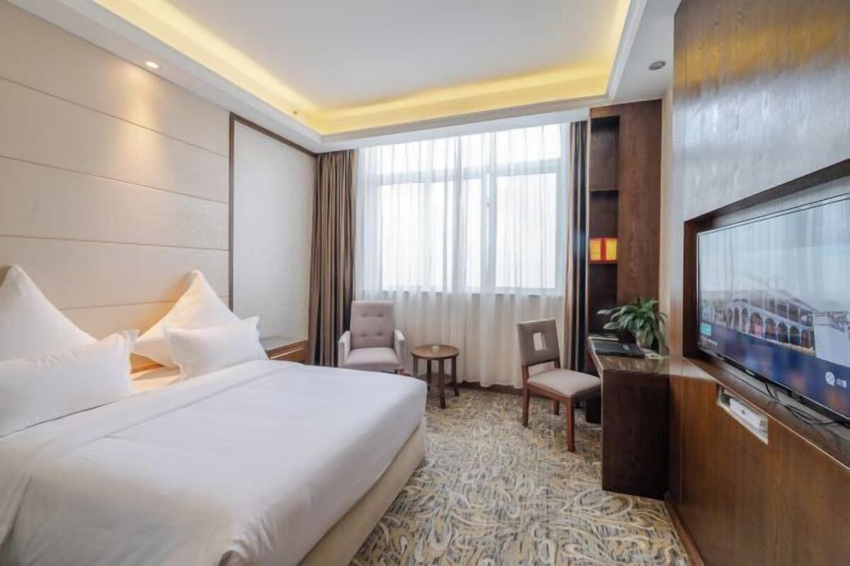 Habitación de hotel con cama, dos sillones, una mesita, televisor y un amplio ventanal con cortinas.