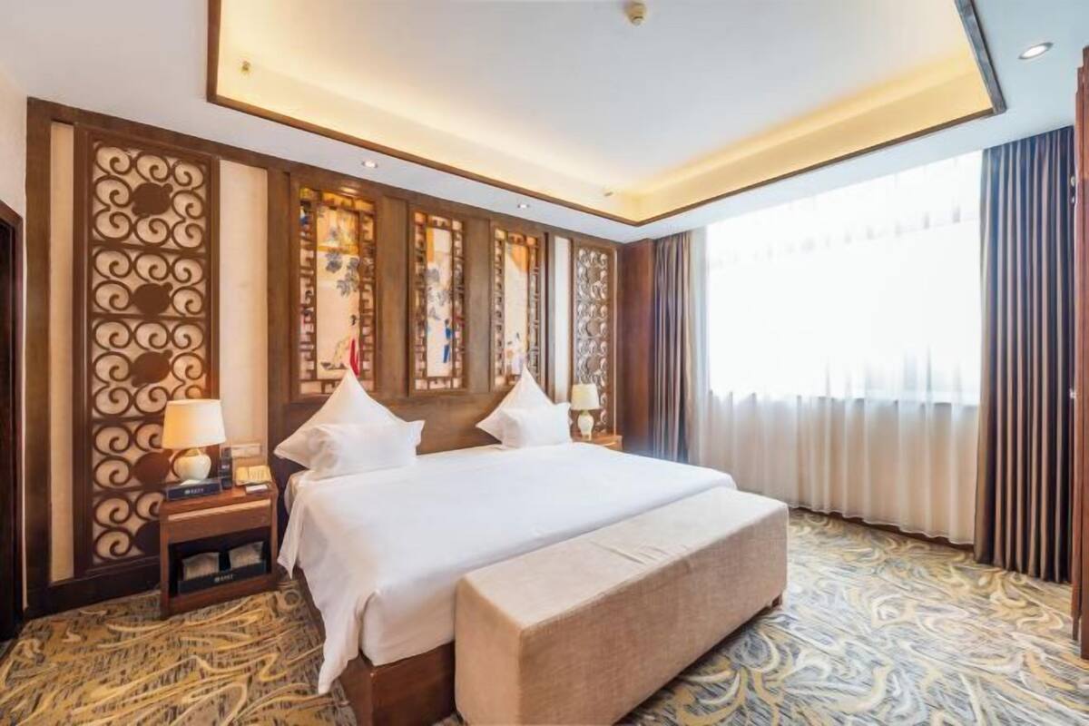 Habitación de hotel con una cama grande, una mesita de noche con lámpara, un panel decorativo en la pared y vistas al exterior.