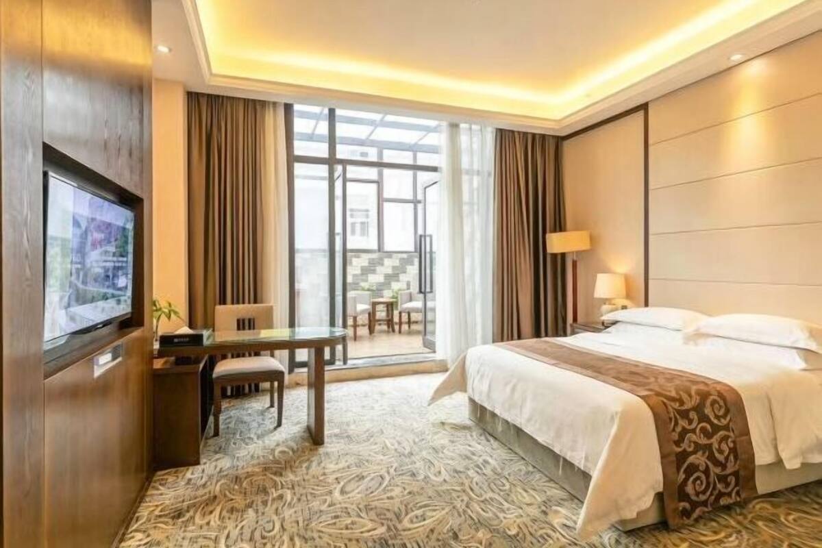 Habitación de hotel con cama, televisor, escritorio con silla, ventana con cortinas y vista al exterior.