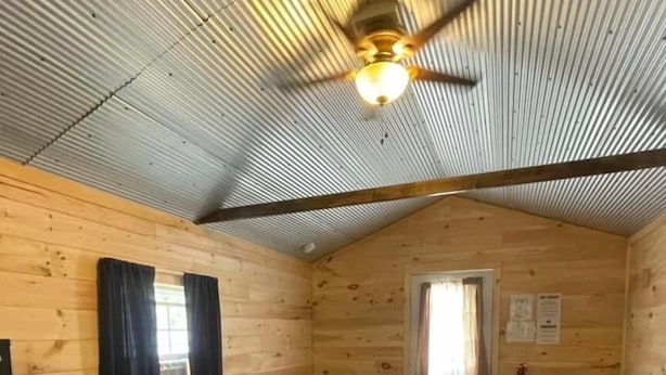 Un dormitorio rústico de cabaña de madera con una cama, un ventilador de techo y una ventana con cortinas.