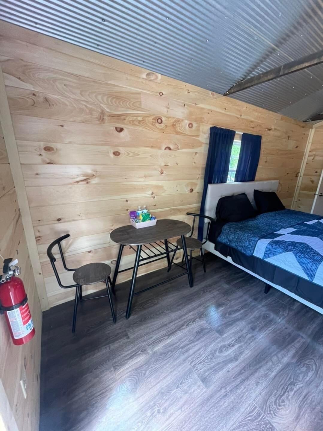 A compact room with a bed, a small table, two chairs, a fire extinguisher, and a window with curtains.