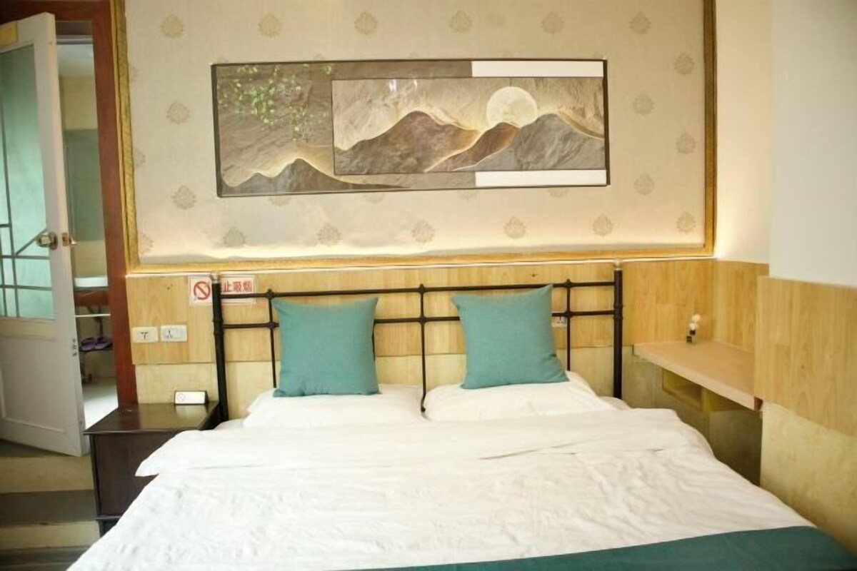 A hotel room with a bed, two pillows, a nightstand, and a wall-mounted artwork depicting a mountain scene.