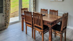 A dining area with a wooden table and chairs, a view of a green field through a glass door, and a framed painting on the wall. - Kingfisher Lodge - UK40017 (Halwill Junction, near Beaworthy)