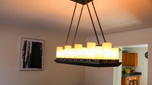 A dining area with a chandelier, a framed abstract painting, and a kitchen visible in the background.