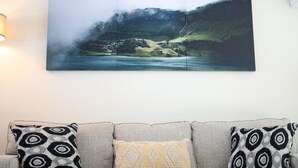 A living room with a large landscape painting on the wall, a sofa with patterned cushions, and a lamp on the left.