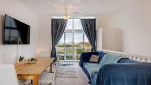 A room with a wooden table, a blue sofa, a TV mounted on the wall, and a view through a large window. - 2 bedroom accommodation in Hunstanton (Hunstanton)