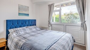 A bedroom with a blue headboard, a plaid bedspread, a small bedside table, a window with curtains, and a painting on the wall. - 2 bedroom accommodation in Hunstanton (Hunstanton)
