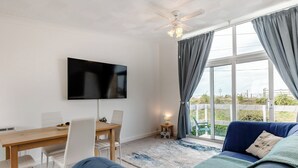 A modern living room with a flat-screen TV, a ceiling fan, a dining table with chairs, and a large window with curtains. - 2 bedroom accommodation in Hunstanton (Hunstanton)