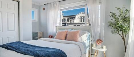 A bedroom with a bed, a window with curtains, a bedside table with a vase, and a view of buildings outside.
