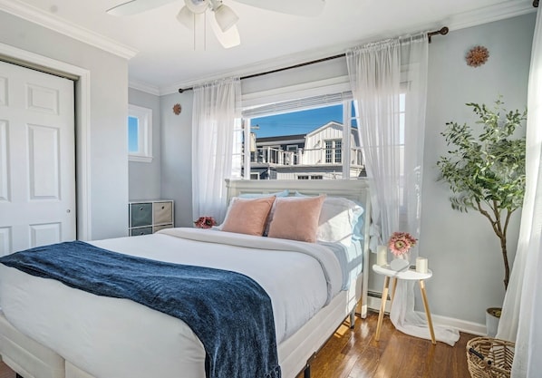 A bedroom with a bed, a window with curtains, a bedside table with a vase, and a view of buildings outside.
