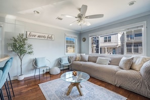 A living room with a sofa, chairs, a glass coffee table, a ceiling fan, and a wall with a sign.