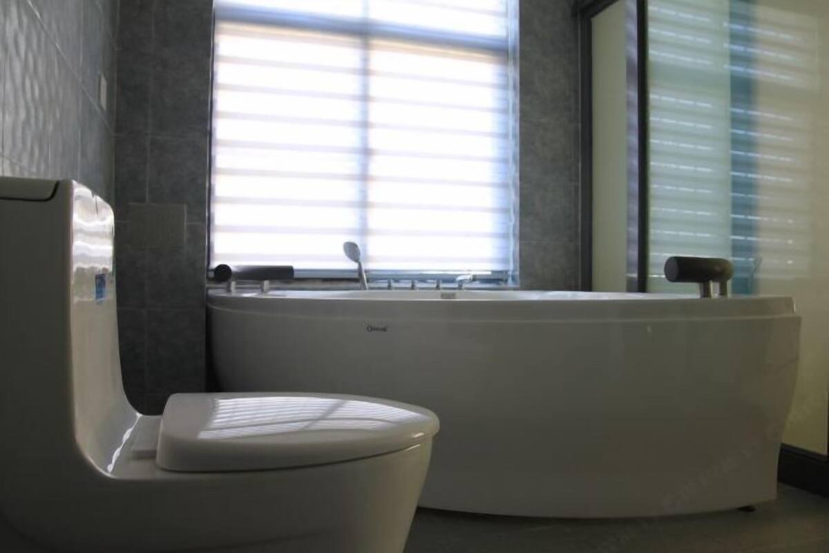 A modern bathroom with a white toilet, a bathtub, and a window with blinds.