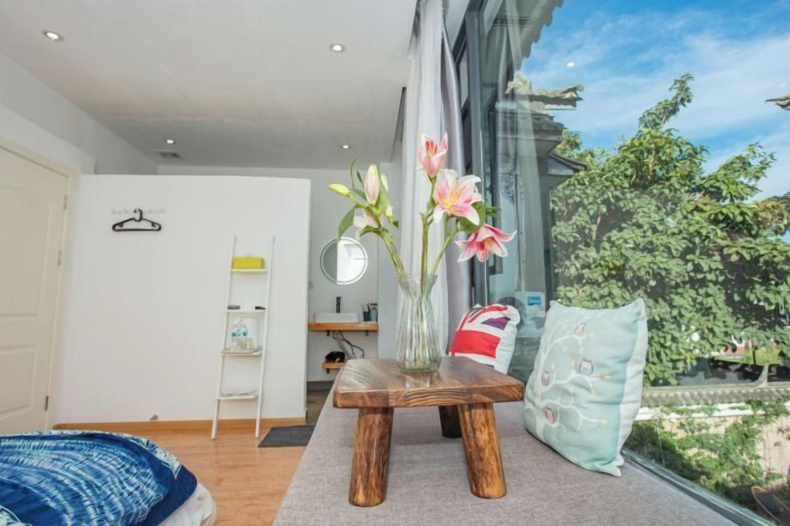 A modern room with a wooden table, a vase with flowers, a window with a view of greenery, and a bed with a blue cover.